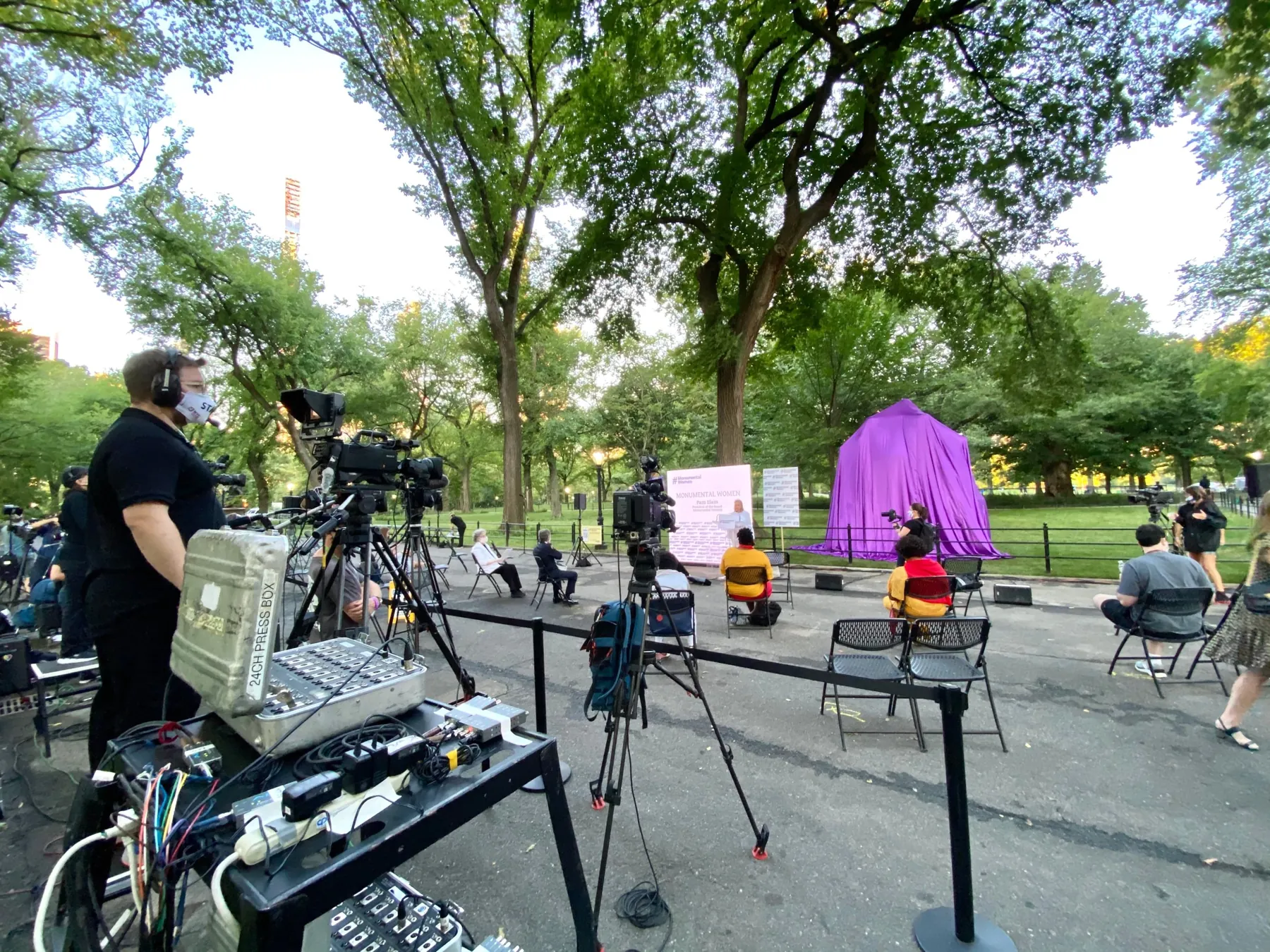 Broadcast cameras and a press box at the Monumental Women unveiling in Central Park, with the covered statue and socially distanced guests.