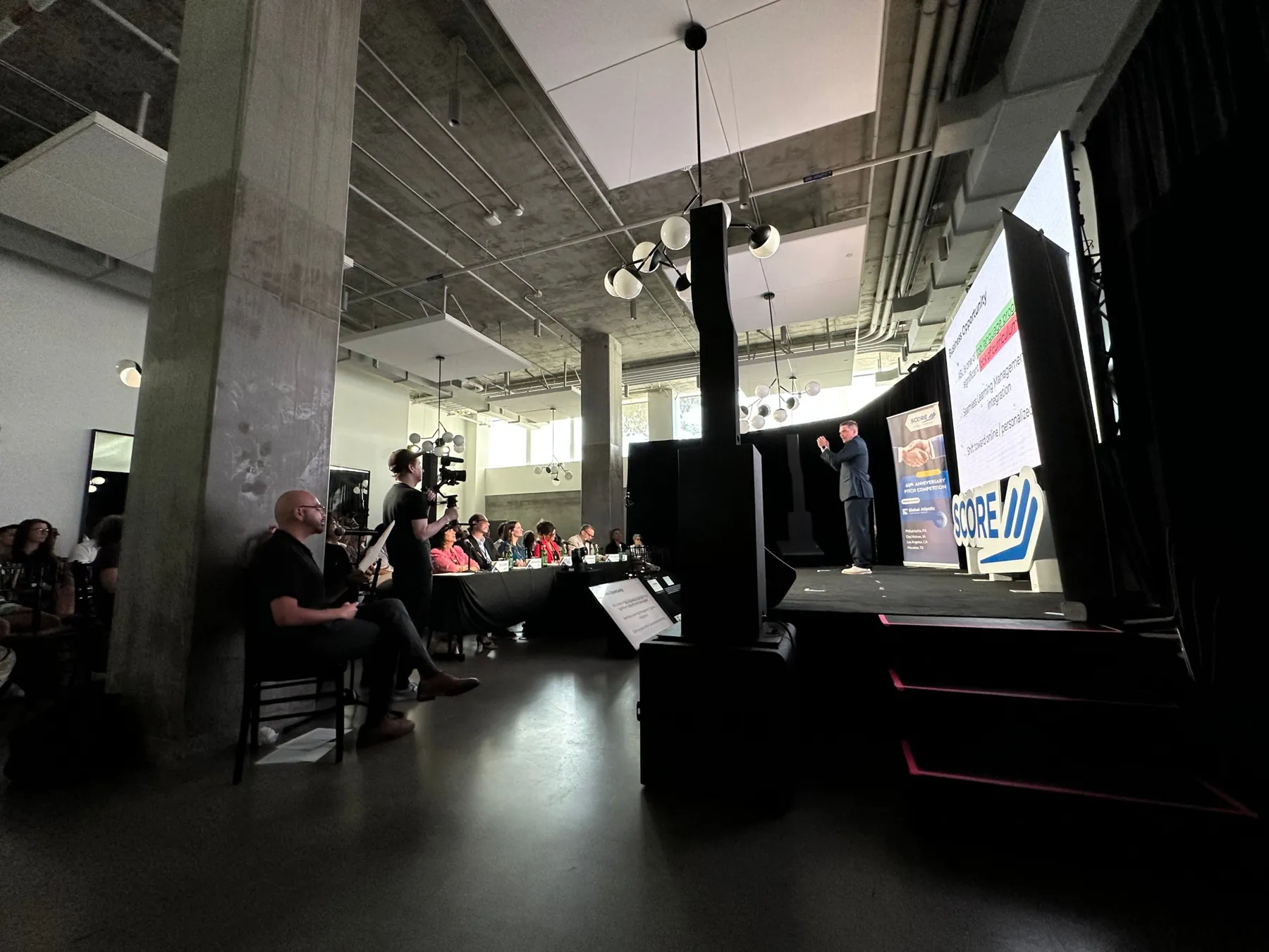 An entrepreneur pitches on stage while the judges' table and a camera operator look on at SCORE's 60th Anniversary Pitch Competition.