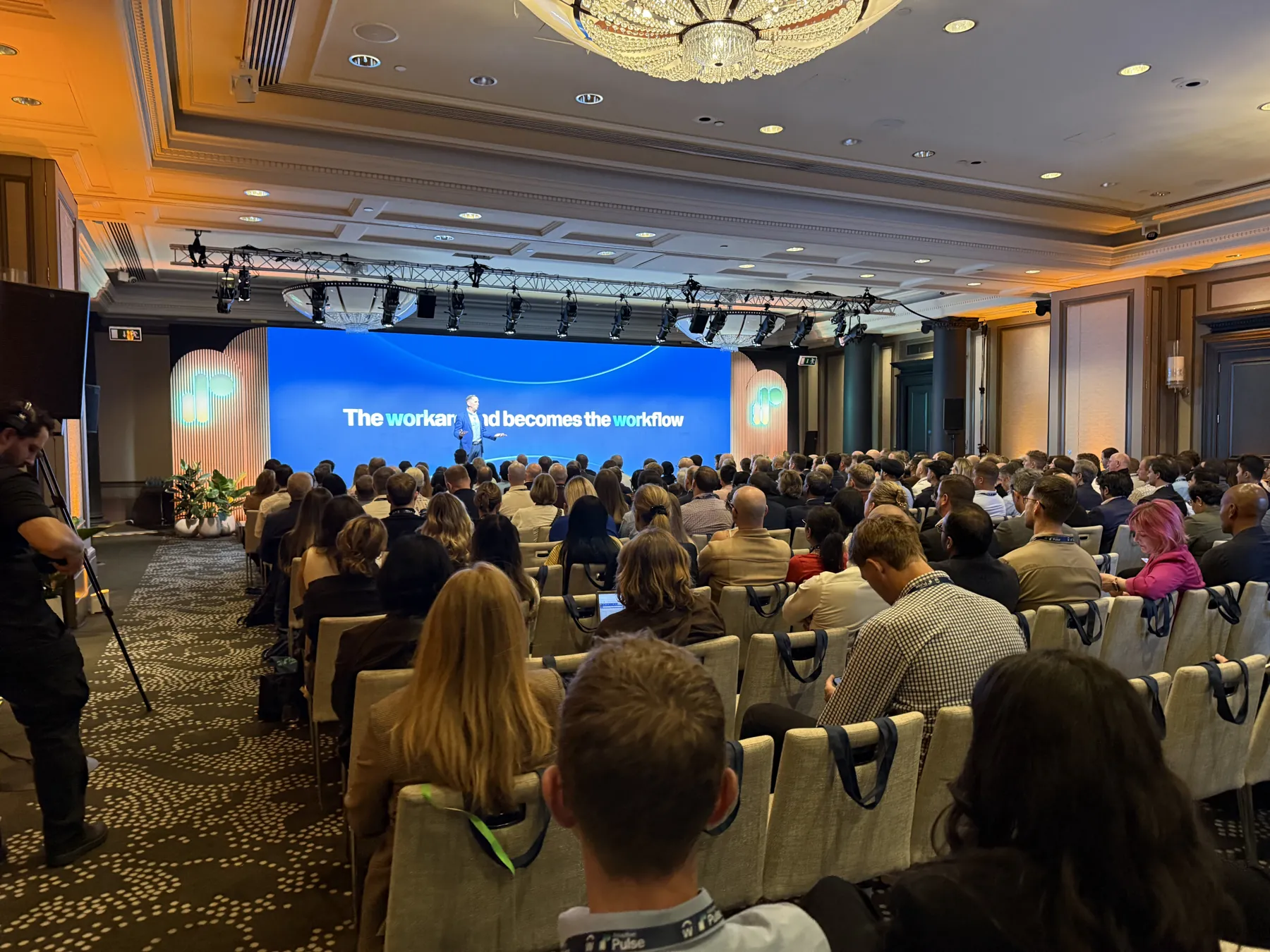 A full hotel ballroom watching a presenter in front of a wide blue LED wall.