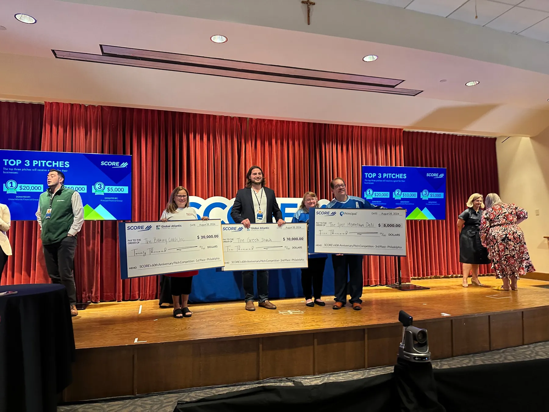 Three winners hold oversized checks on stage at SCORE's 60th Anniversary Pitch Competition in Philadelphia.