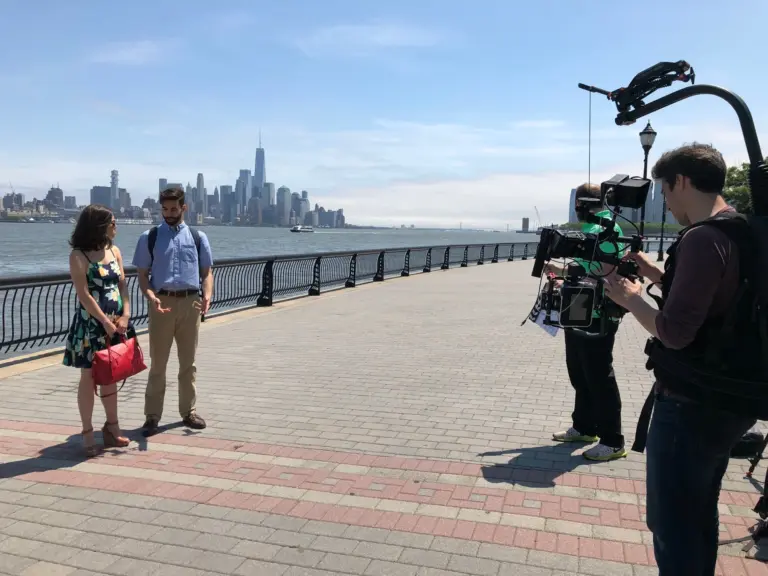 A two-person interview shoot on a waterfront walkway, with a camera operator on a stabilizer rig and the lower Manhattan skyline across the river.