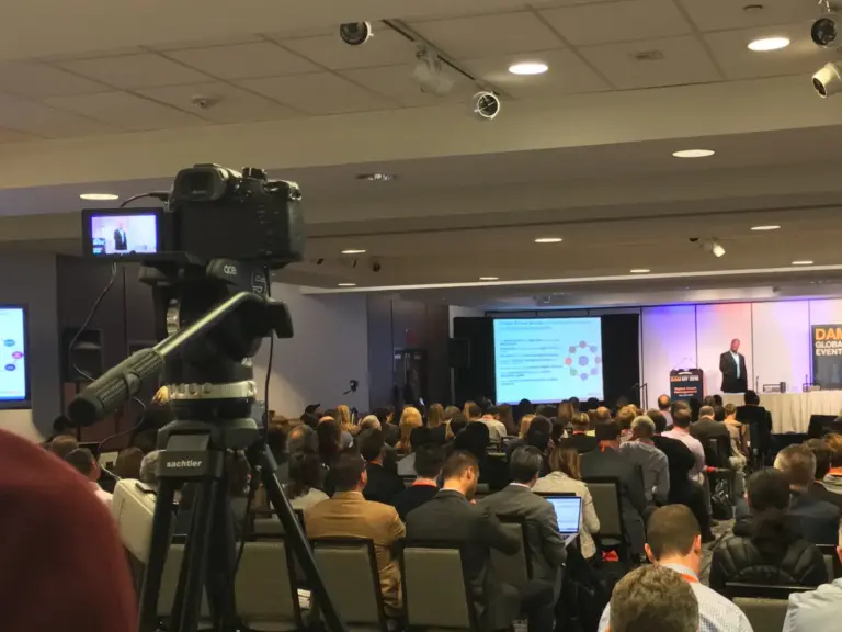 A camera on a tripod at the back of a packed conference session, with a presenter on stage and slides on screen.