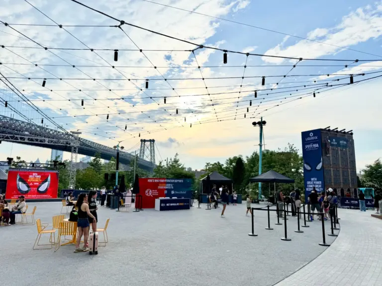 An outdoor brand activation in Domino Park, Brooklyn, under a string-light canopy, with an LED wall, branded climbing wall and guest lines, the Williamsburg Bridge behind.