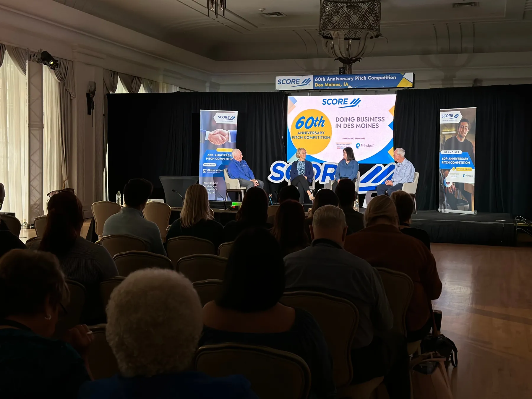 A four-person panel on stage at SCORE's 60th Anniversary Pitch Competition in Des Moines, in front of a seated audience.