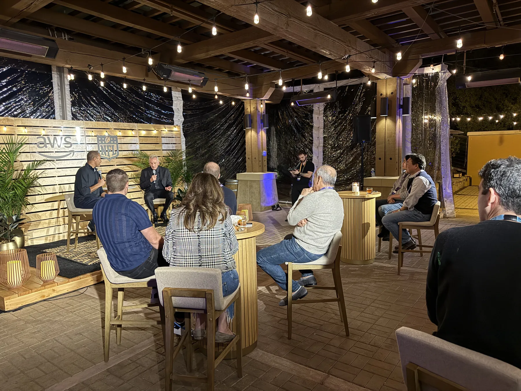 A fireside chat on an outdoor stage under string lights, with guests seated at high-top tables.