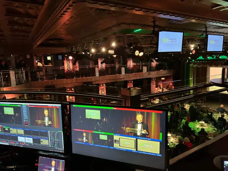 View from the balcony show desk at a gala: switcher monitors in front, an honoree at the podium on screen and dinner tables below.