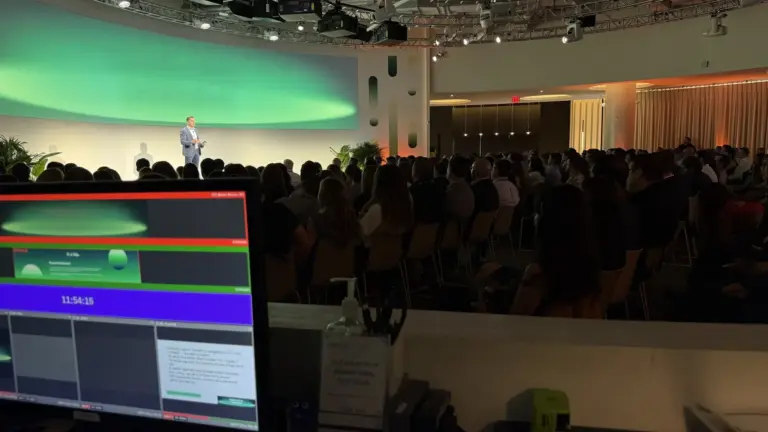 A presenter on stage beneath a green LED wall, seen past a show-desk monitor, with a packed audience.
