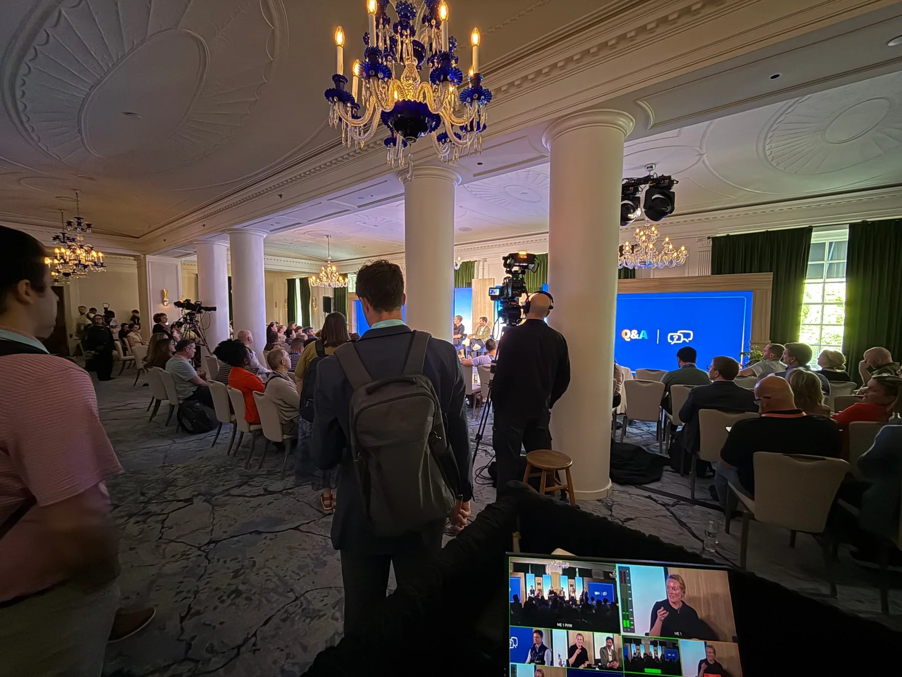 A conference panel in a hotel ballroom seen from the production position, with a camera operator on headset, a Q&A slide on the LED wall and the live switcher multiview in the foreground.