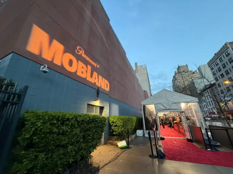 Premiere red carpet at dusk: a gobo-projected title on the building wall and a tented carpet with press lines and barricades.
