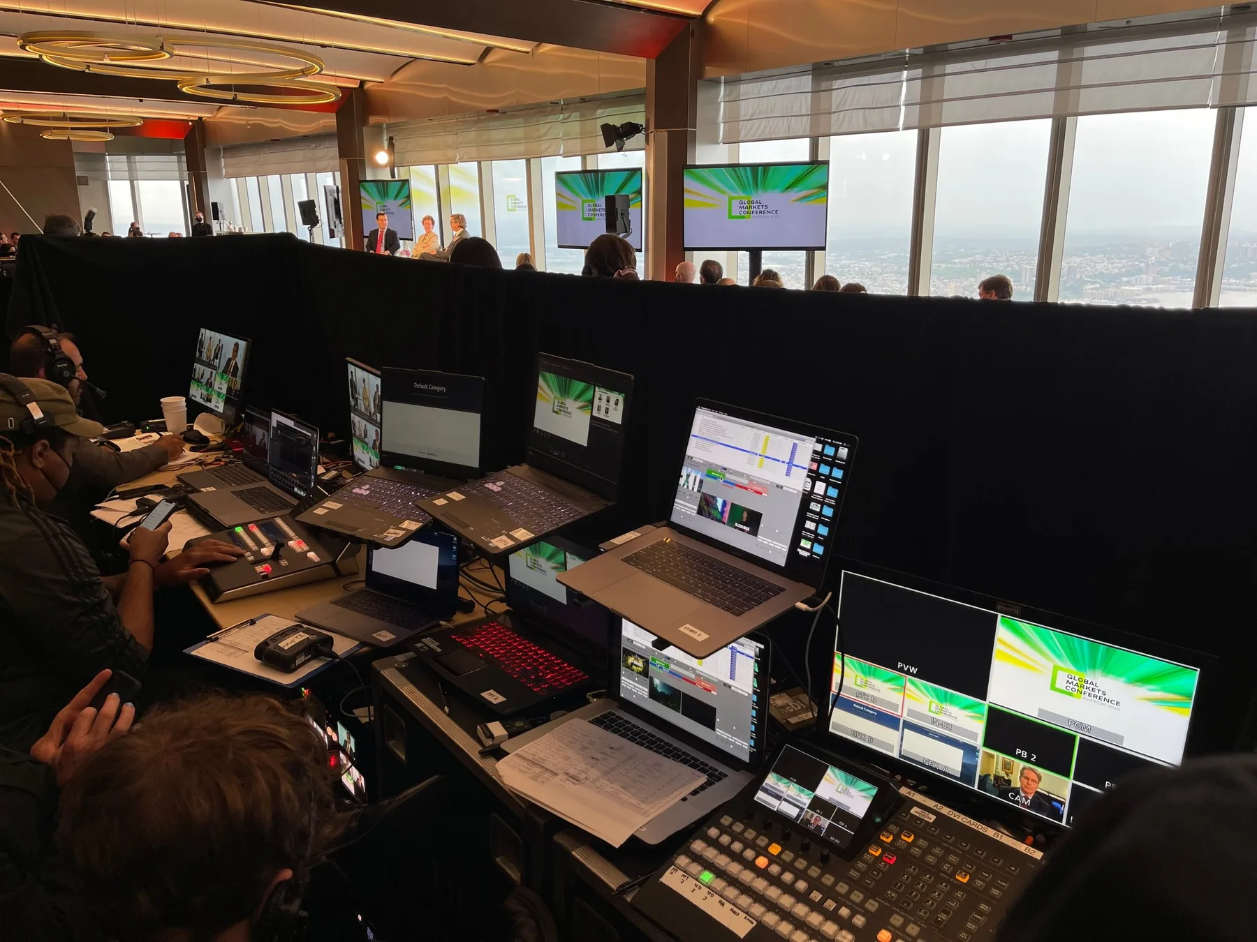 Our production position at a financial conference high above the Hudson: switcher, graphics and playback stations behind pipe and drape, with a panel on stage beyond.